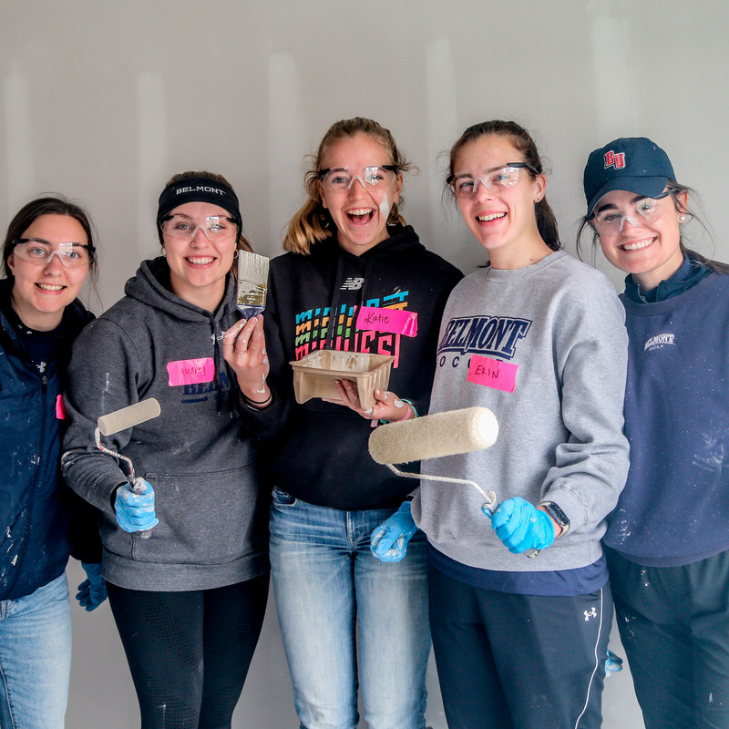 Five students holding rollers at a Habitat build site wearing sweatshirts and safety goggles