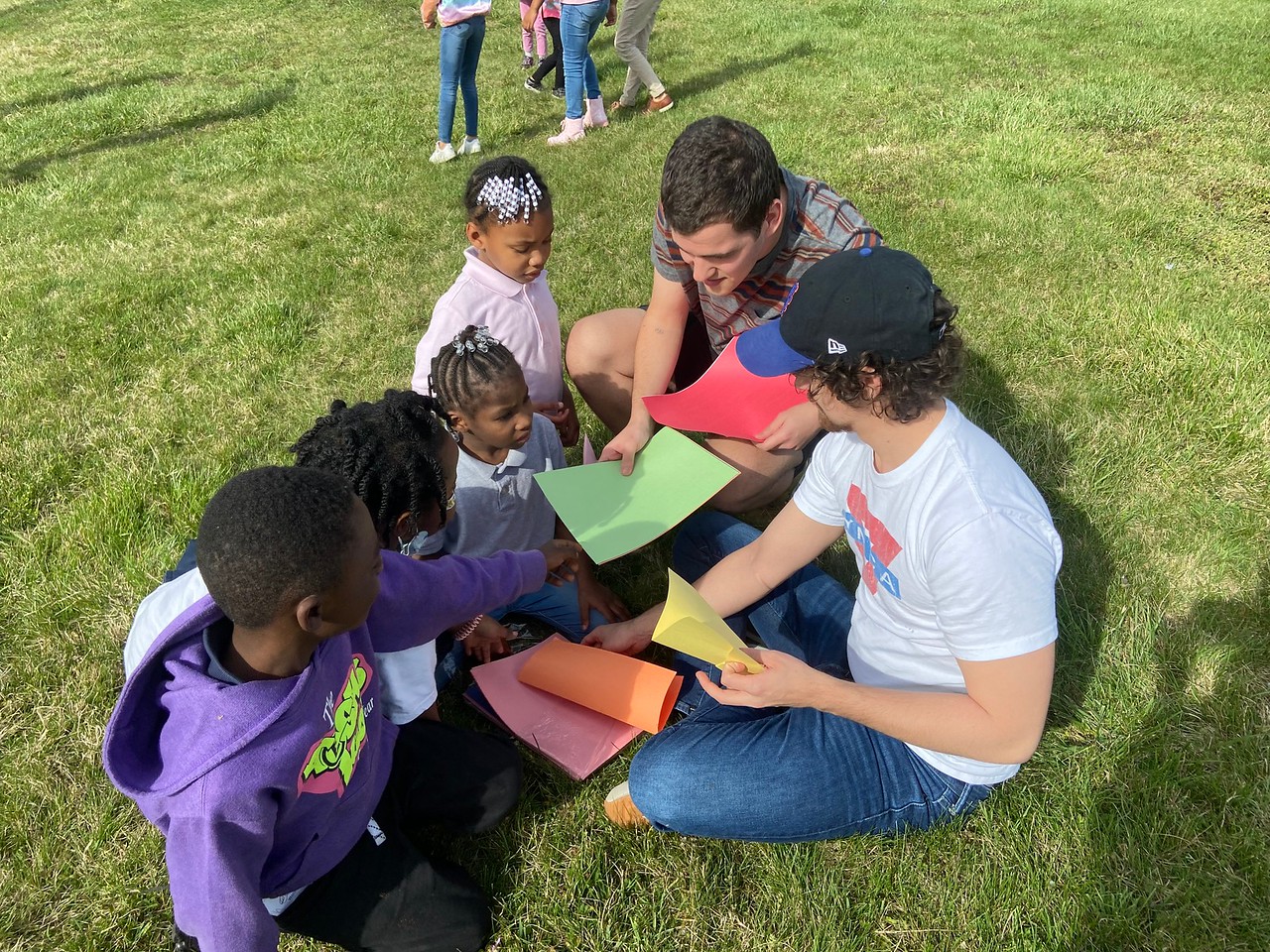 students handing out colored paper for four children while sitting in the grass