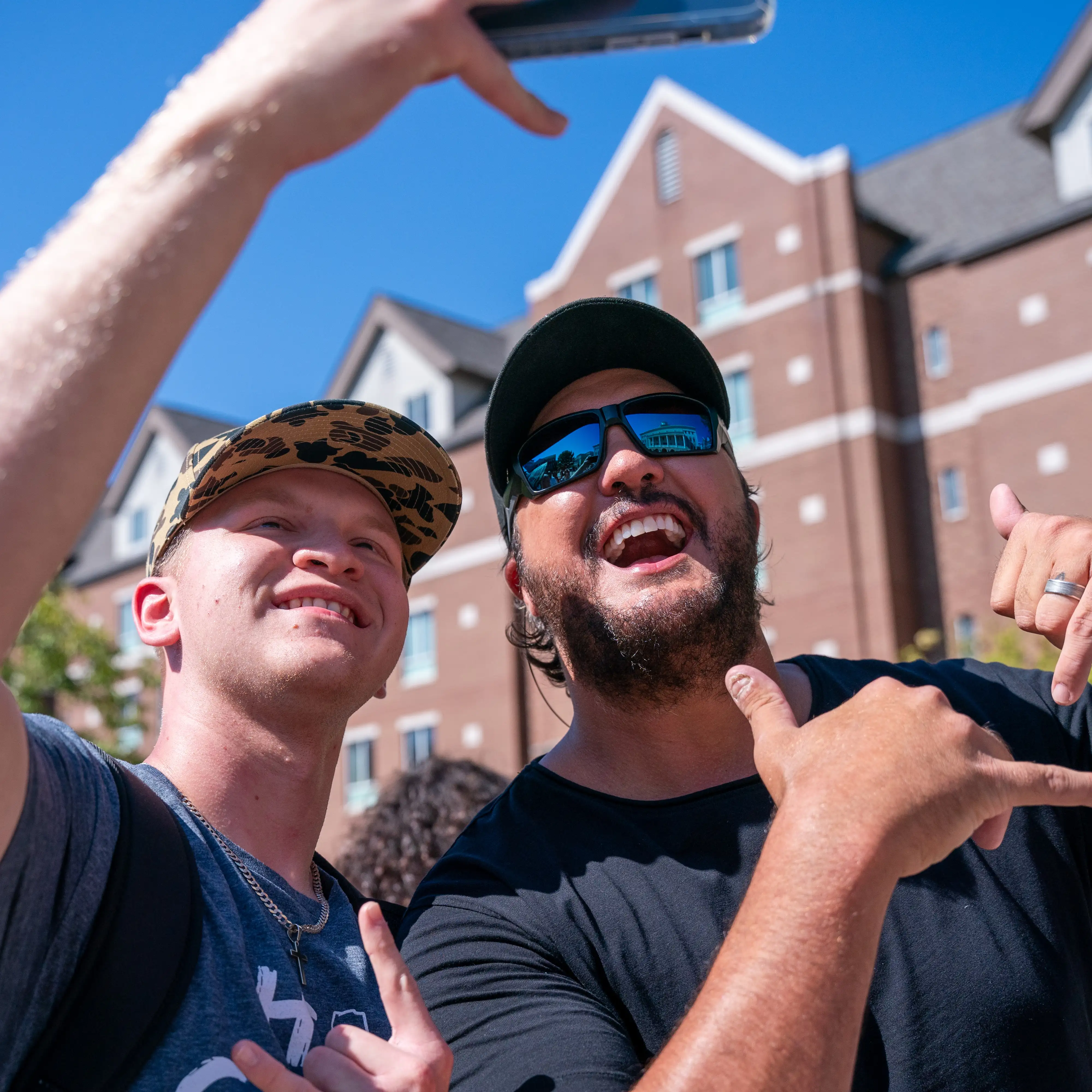 Two smiling men pose for a selfie outdoors in front of a brick building with white-trimmed windows. The man on the left wears a camo-patterned baseball cap, a gray T-shirt, and a silver chain necklace; he holds up a phone to take the photo. Country artist Luke Bryan on the right wears a black T-shirt, black cap, and mirrored sunglasses, sports a short beard, and makes a “hang loose” hand gesture with his right hand.