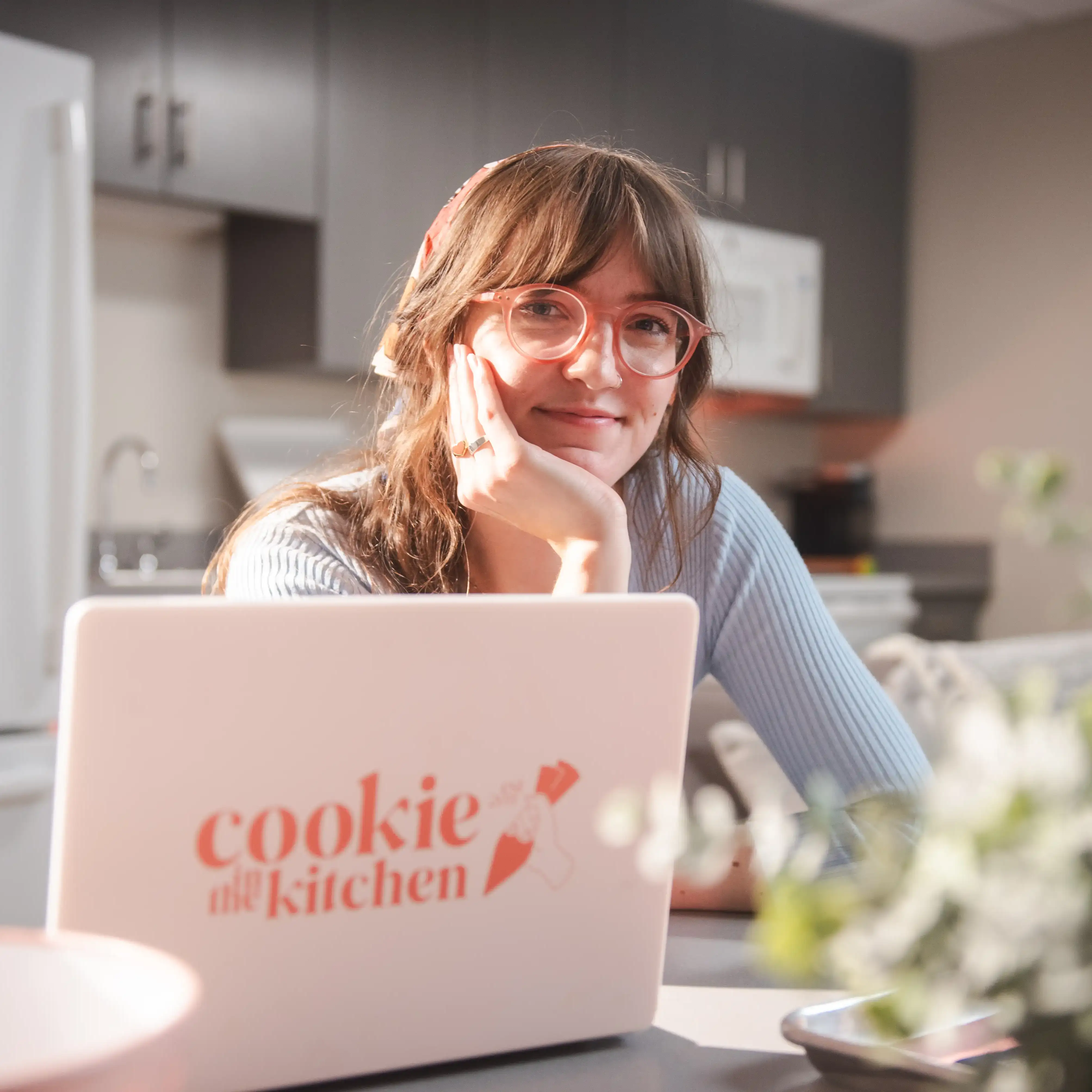 A smiling young woman with long brown hair, pink-framed glasses, and a headband rests her chin on her hand as she leans over a white laptop displaying a red “cookie the kitchen” logo. She sits in a modern kitchen setting with blurred cabinets, a sink, and a stovetop in the background.