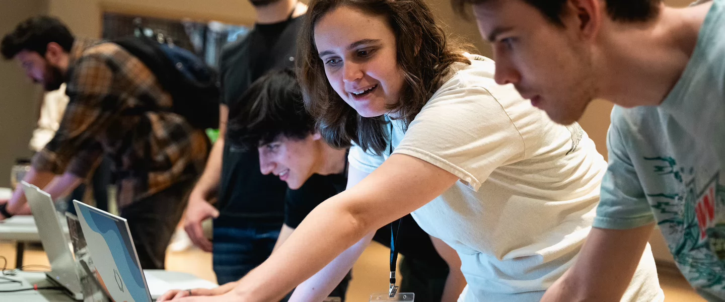 A group of young adults enthusiastically interact with laptops on a table. The room's atmosphere is energetic and collaborative, suggesting a workshop or hackathon.