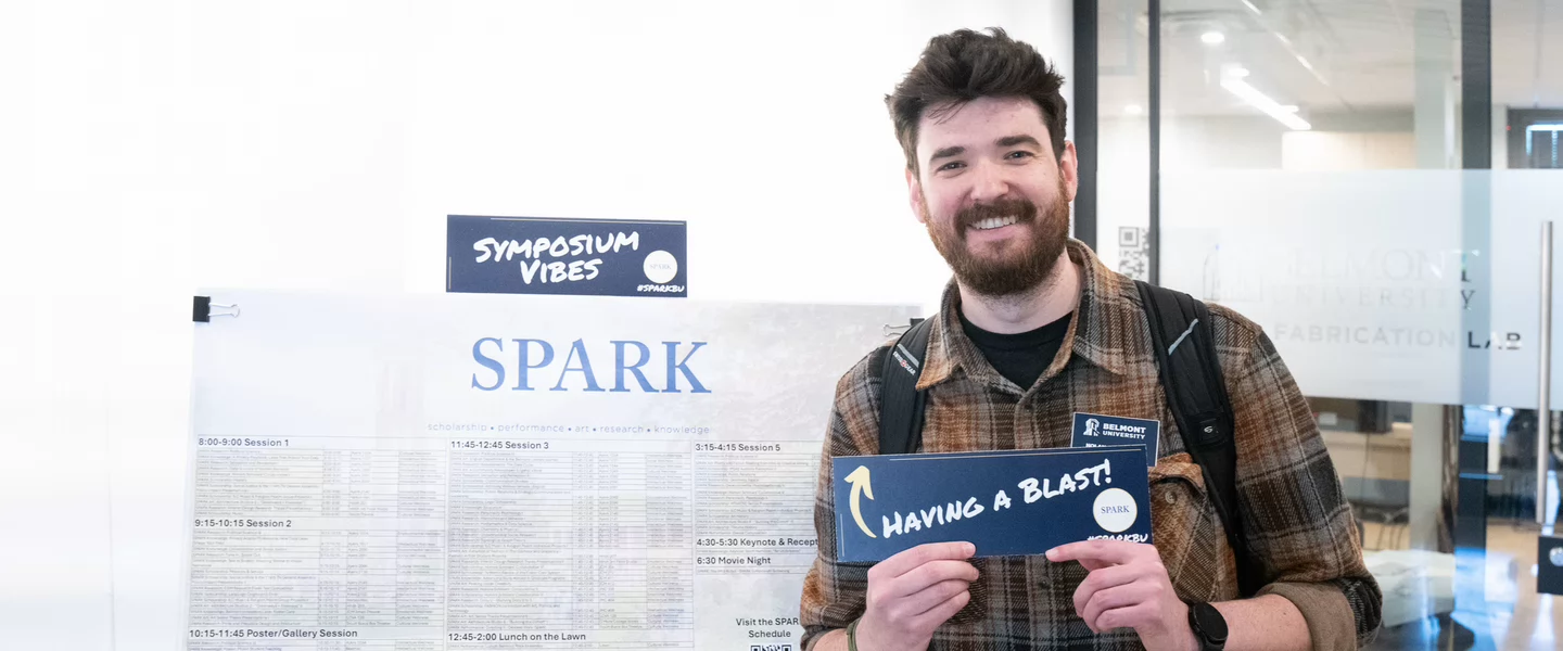 Man holds a "Having a Blast" sign at SPARK symposium. He stands beside a schedule marked "SPARK." 