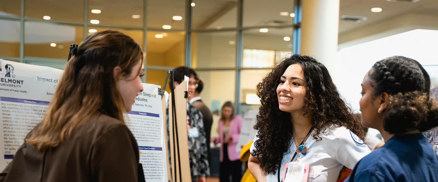 A young woman in a white coat smiles warmly while talking to colleagues near a research poster in a bright conference room.