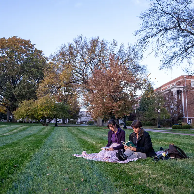 Two young women are sitting on a blanket in a grassy area surrounded by trees with autumn foliage. One woman is reading a book while the other is writing in a notebook. They are dressed warmly, reflecting the cool weather. In the background, a large building is partially visible, and the sky is clear with a hint of evening light.