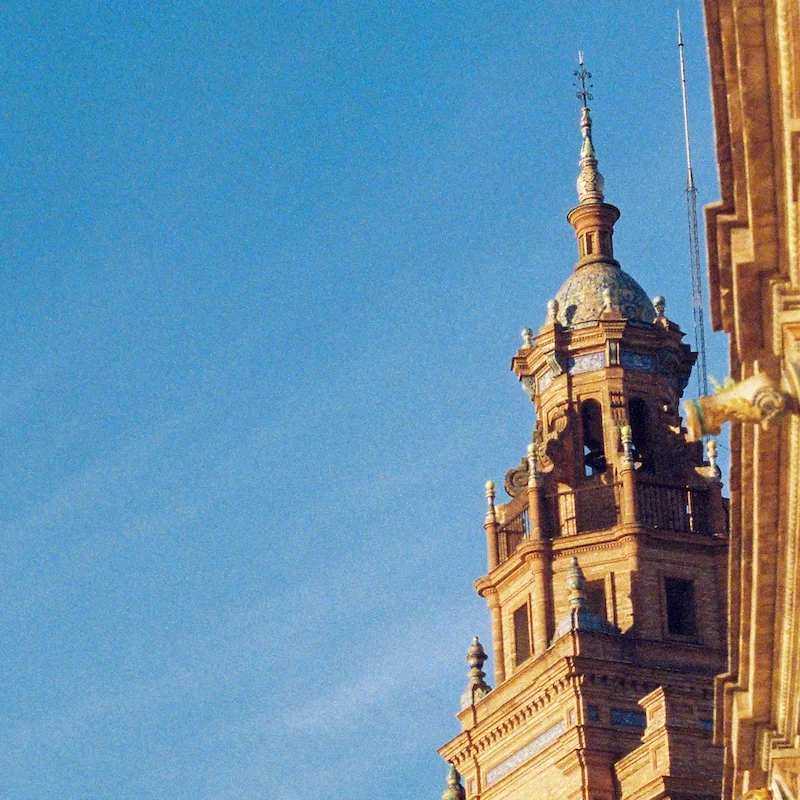 Close-up view of an ornate cathedral bell tower in Seville, Spain, featuring Baroque architecture with a decorative dome topped by a spire. The intricate stonework appears golden in the sunlight against a clear blue sky. Part of another building structure is visible on the right edge of the frame.