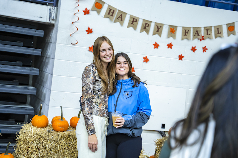 Two student pose in front of fall festival backdrop