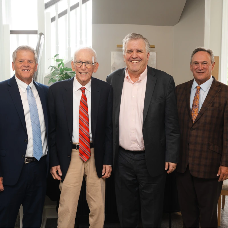 Four smiling businessmen in suits, diverse ages, standing together in a modern office environment.