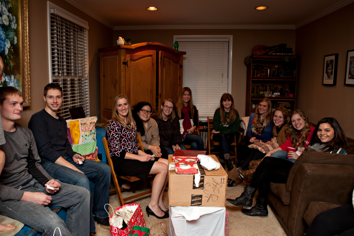 Group of young adults smiling in a living room surrounded by gifts, likely during a holiday celebration or party.