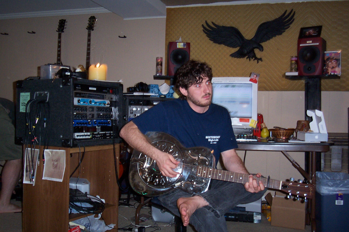 Man with curly hair playing a chrome resonator guitar in a home music studio with audio equipment and wall-mounted guitars.