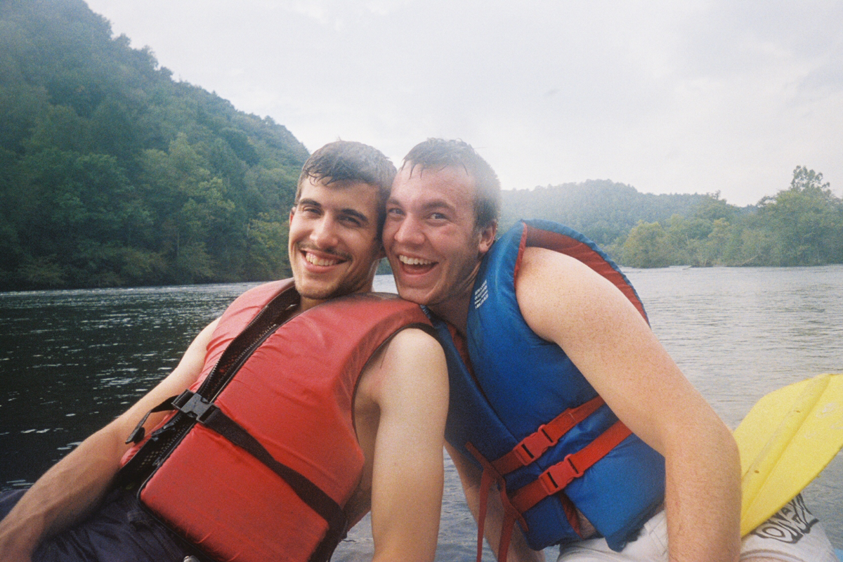 Two smiling men in red and blue life jackets boating on a river with green mountains.