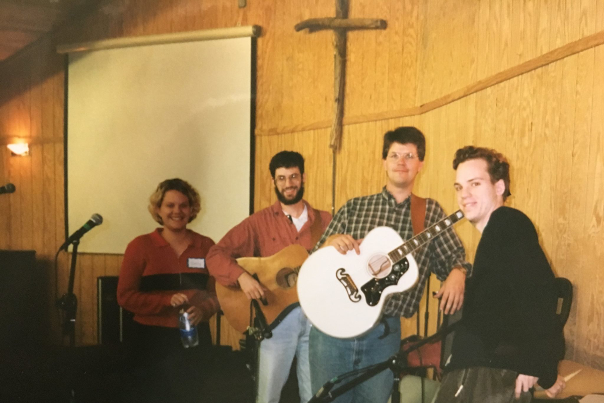 Music group of four, two playing acoustic guitars, smiling in a venue with a crucifix and screen.