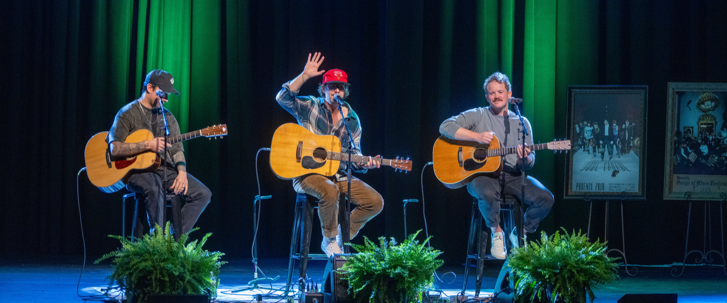 Acoustic trio performing live on stage, playing guitars under green lights, with one musician waving.