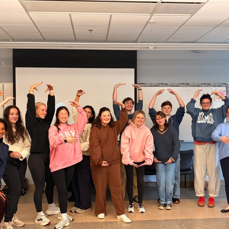 A group of approximately a dozen young students stand together on a classroom stage with their arms raised in celebration, wearing casual clothing including hoodies and sweaters. A projection screen is visible in the background, with fluorescent ceiling lights overhead.