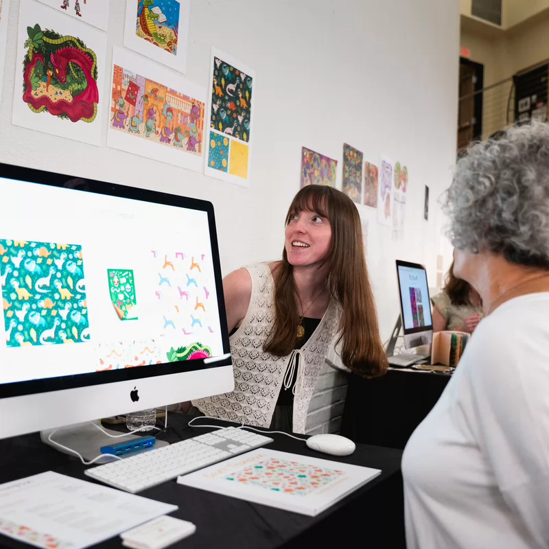 A woman with long brown hair enthusiastically presents colorful digital designs on a computer to an older woman with curly gray hair at Watkins Art Crawl.