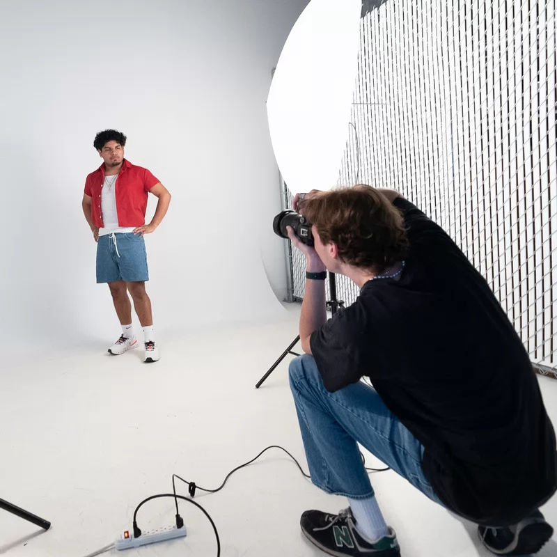 Photographer crouches, taking photos of a standing model in a casual outfit. Bright studio lights illuminate the scene, creating a focused atmosphere.