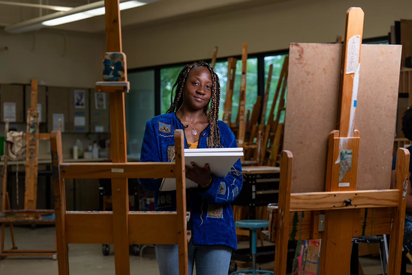 Woman in a blue sweater holds a sketchbook in an art studio filled with wooden easels. She appears focused and content, with a creative atmosphere.