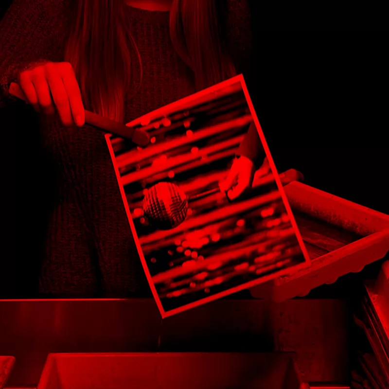 A person develops a black-and-white photo in a darkroom under red lighting. The image of a disco ball emerges in a tray of liquid, evoking a focus on photography processes.